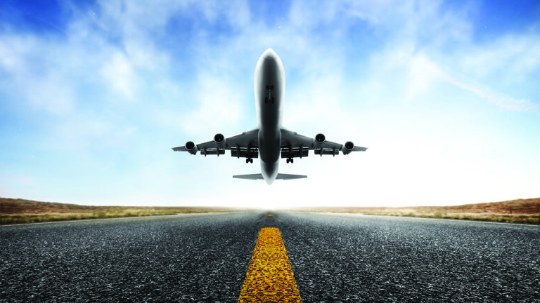 An airplane takes off from a runway with a clear blue sky in the background. The image is captured from a low angle directly in front of the plane.