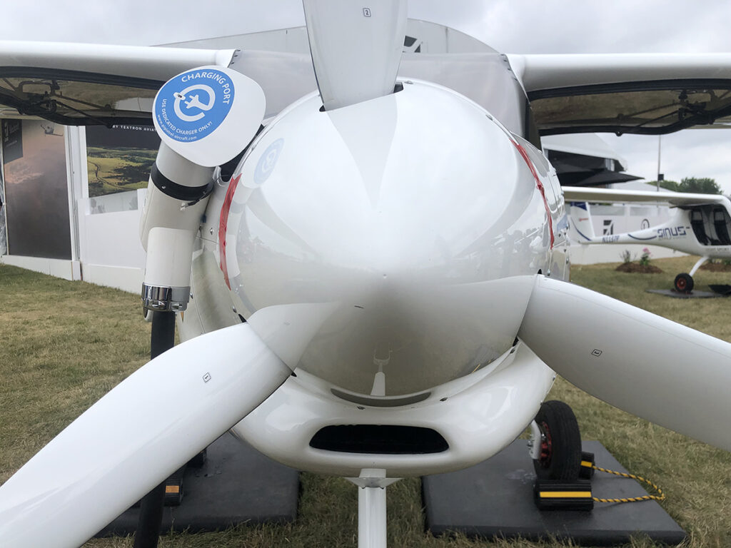 Close-up view of the nose and propeller of a small white aircraft, with a charging port labeled "charging" visible on the left side. The aircraft is parked on grass near an exhibition booth.