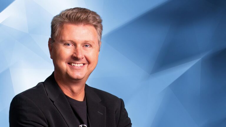 A person with short, light brown hair and wearing a black blazer stands in front of a blue geometric background, smiling at the camera.