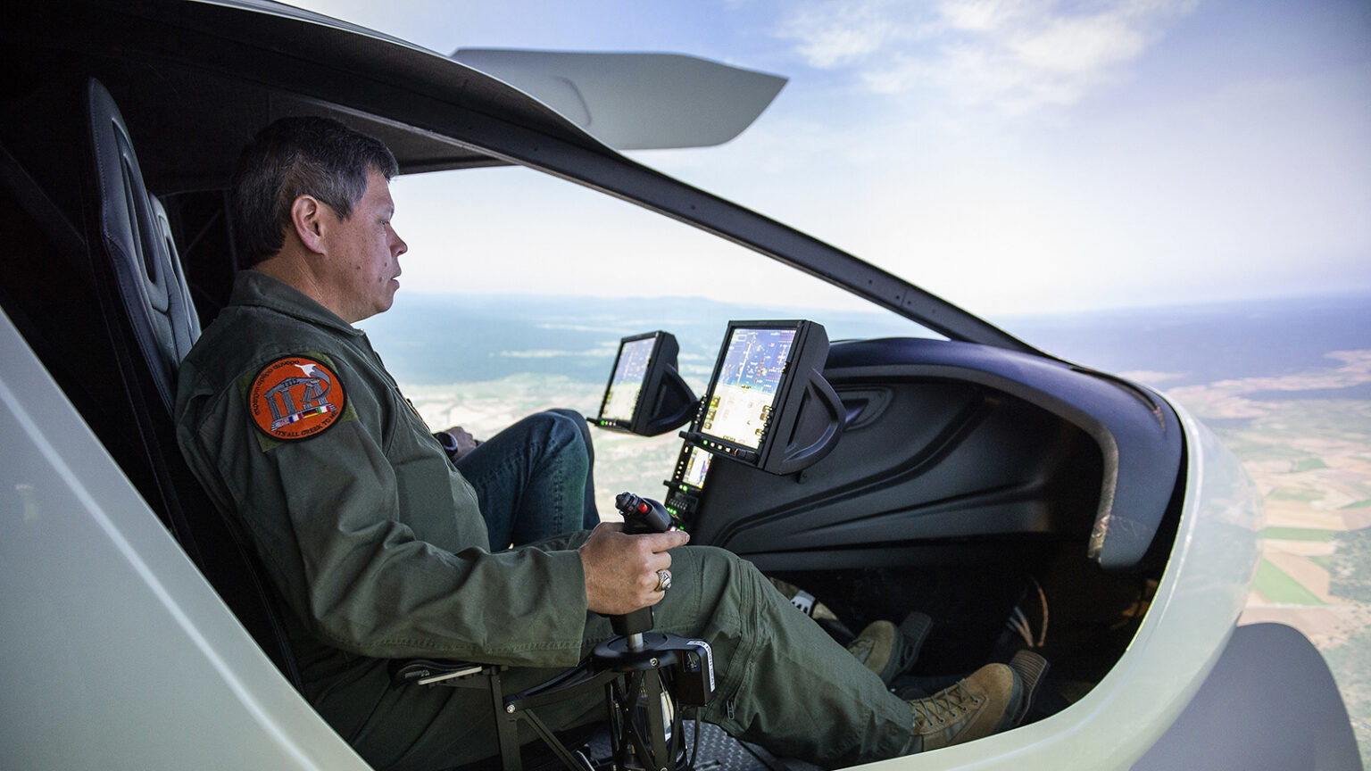 A person operating a flight simulator with multiple screens inside a cockpit-like setup, wearing a green flight suit and gripping a joystick.