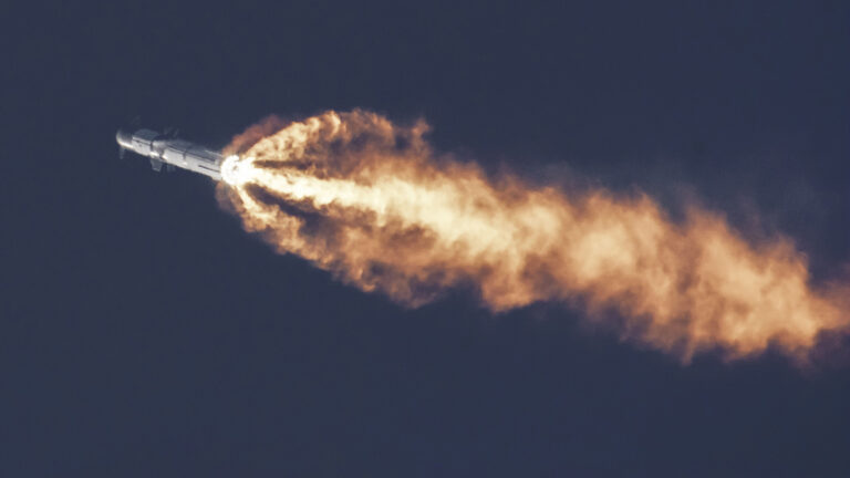 A rocket is captured during liftoff, with bright orange flames trailing from its engines against a dark blue sky.