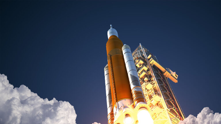 A large rocket is launching into the sky, with flames and smoke at its base. The rocket is orange and white, and it is partially framed by a metal launch tower against a clear blue sky.
