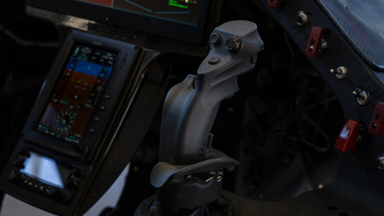 Close-up of an aircraft cockpit showing a central control stick, various buttons, and display screens.