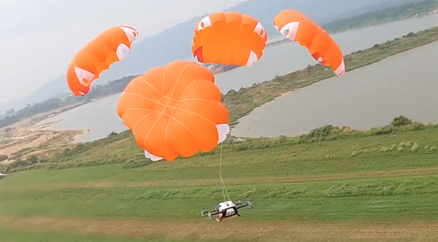 A drone is descending, aided by three orange parachutes, over a grassy area near a body of water with a distant shoreline.
