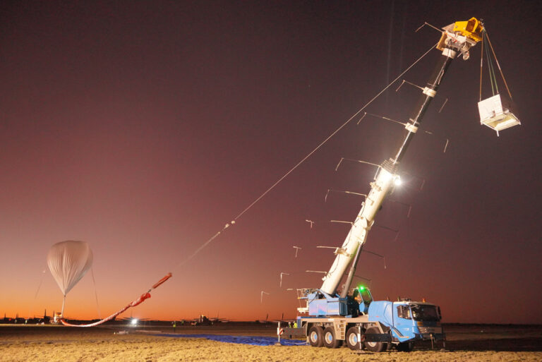A crane hoists a scientific payload attached to a high-altitude balloon at dusk. The balloon is partially inflated, and a truck is positioned next to the crane on an outdoor site.