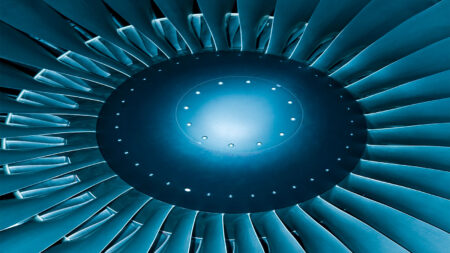Close-up of an aircraft jet engine turbine, showing the circular fan blades and central hub, illuminated with a blue tint.