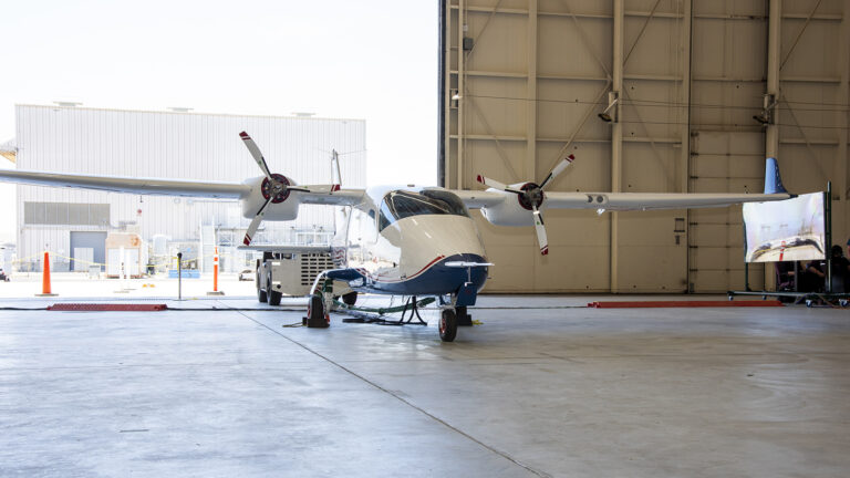 A small twin-propeller aircraft is stationed on the concrete floor of a hangar, with both hangar doors open allowing sunlight to enter.