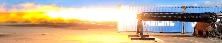 A rocket engine undergoing a test firing, mounted horizontally on a support structure, with a large flame and exhaust plume extending from the nozzle.