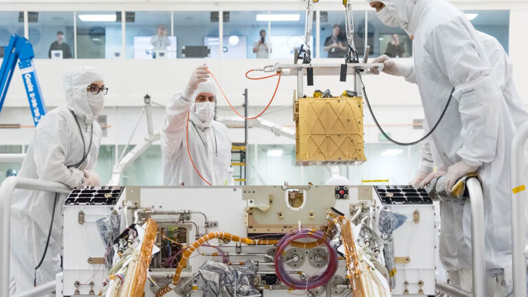 Three people in cleanroom suits work on assembling or inspecting a spacecraft component inside a laboratory, with additional personnel observing from a glass-enclosed area in the background.