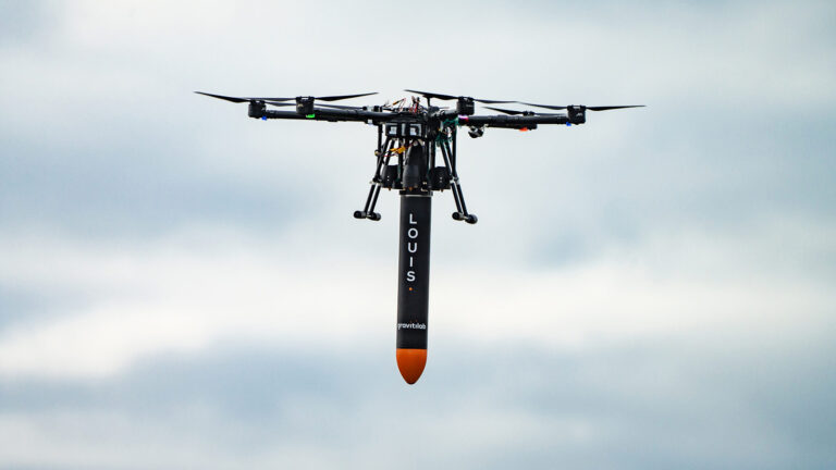 A drone in flight carrying a cylindrical payload labeled "LOUIS." The sky in the background is cloudy.