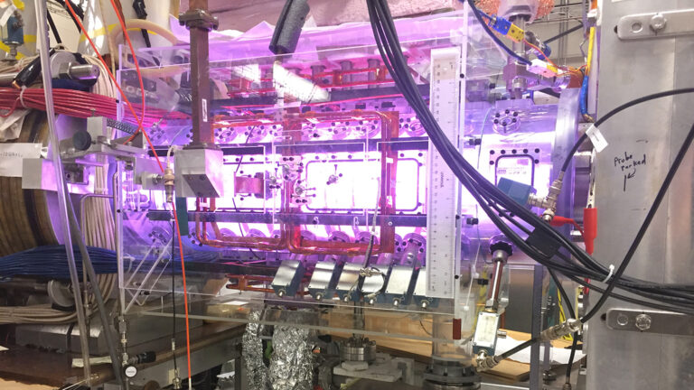 A complex assembly of scientific equipment with numerous wires, tubes, and components, illuminated with a purple light, likely part of a laboratory or experimental setup.