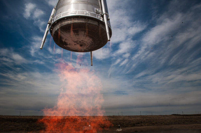A rocket descends gently toward the ground with flames emitting from its engines against a backdrop of a cloudy sky and barren landscape.