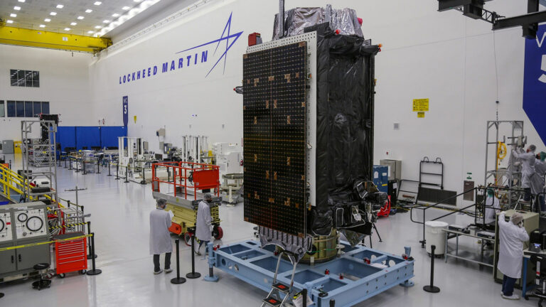 Technicians work on a satellite in a large Lockheed Martin facility.