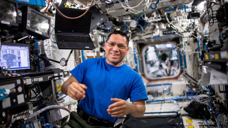 An astronaut in a blue shirt floats in a cluttered space station module, with various equipment and wires around. Another astronaut is visible in the background.