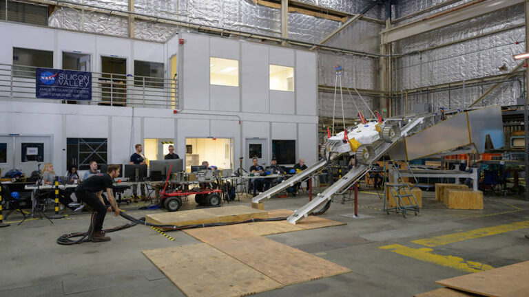A team of engineers monitor and test a Mars rover prototype inside a large industrial facility with metal walls and various equipment. A banner reads "NASA Silicon Valley".