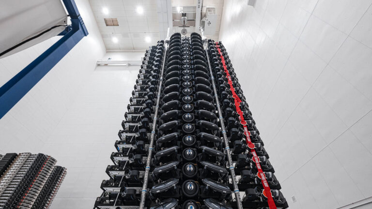 A vertical stack of numerous black drones is shown secured in a storage unit within a large, white, industrial interior.