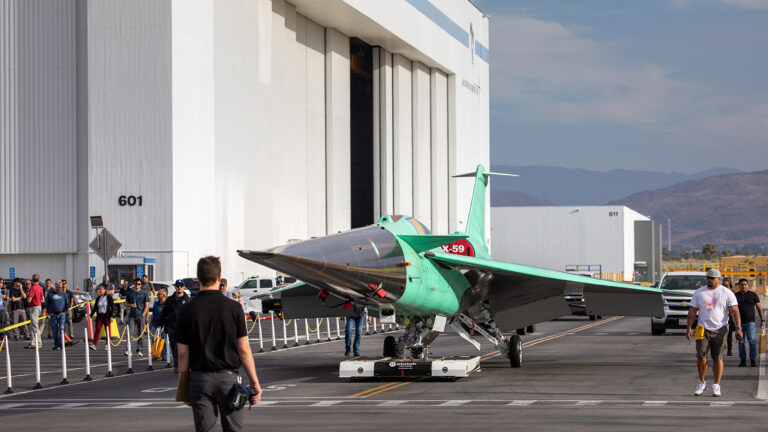 The NASA X-59 supersonic aircraft is moved out of a hangar, surrounded by spectators and workers, against a backdrop of mountains and clear skies.