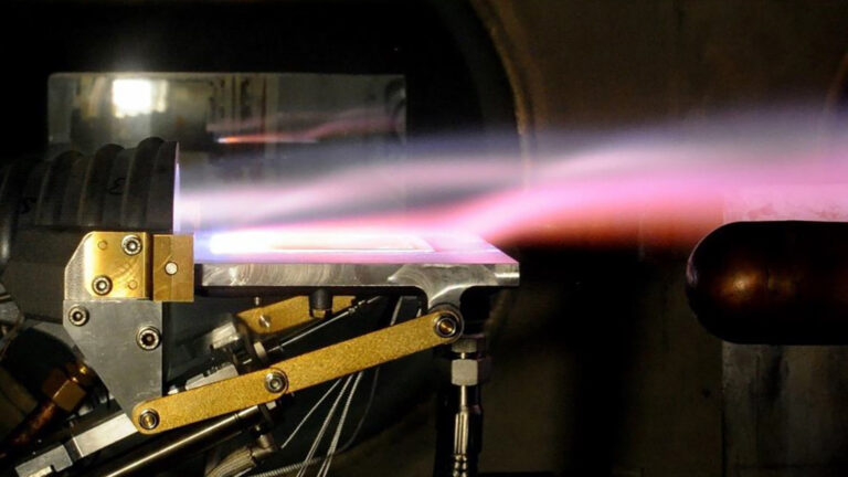 A device undergoing a test, emitting a pale pink and purple flame. The setup includes mechanical components and a metallic surface, indicating a controlled experiment or engineering process.