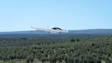 A white electric vertical takeoff and landing (eVTOL) aircraft is flying above a green landscape with clear blue skies in the background.
