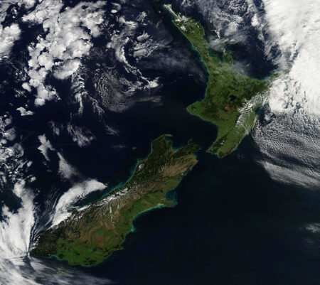A satellite image showing New Zealand's North and South Islands, surrounded by the Pacific Ocean with cloud formations visible to the northwest and southeast.