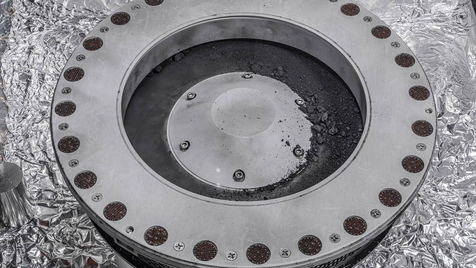 Close-up of a circular metal container with multiple bolts and a central section containing a small amount of dark, granular material, set on a surface covered with crinkled aluminum foil.