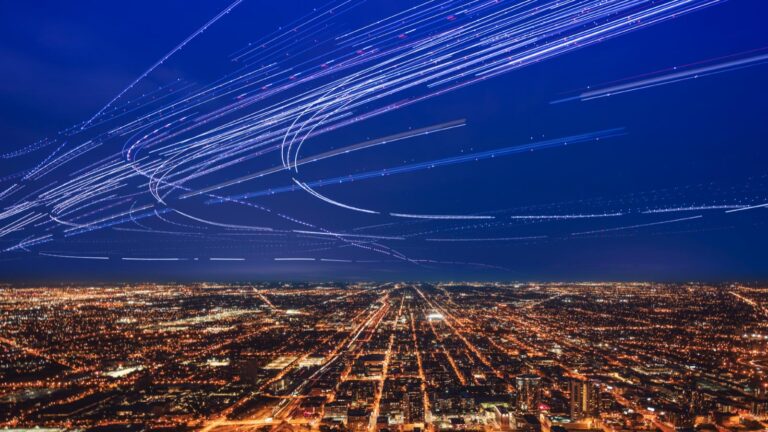 A night cityscape with streaks of light trails from aircraft moving across the sky above the illuminated urban grid below.