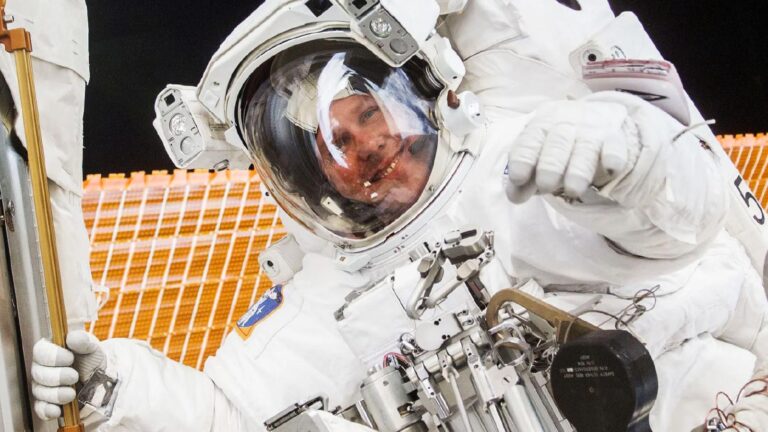 An astronaut in a white spacesuit performs tasks during a spacewalk, with a reflective visor and mechanical equipment visible.