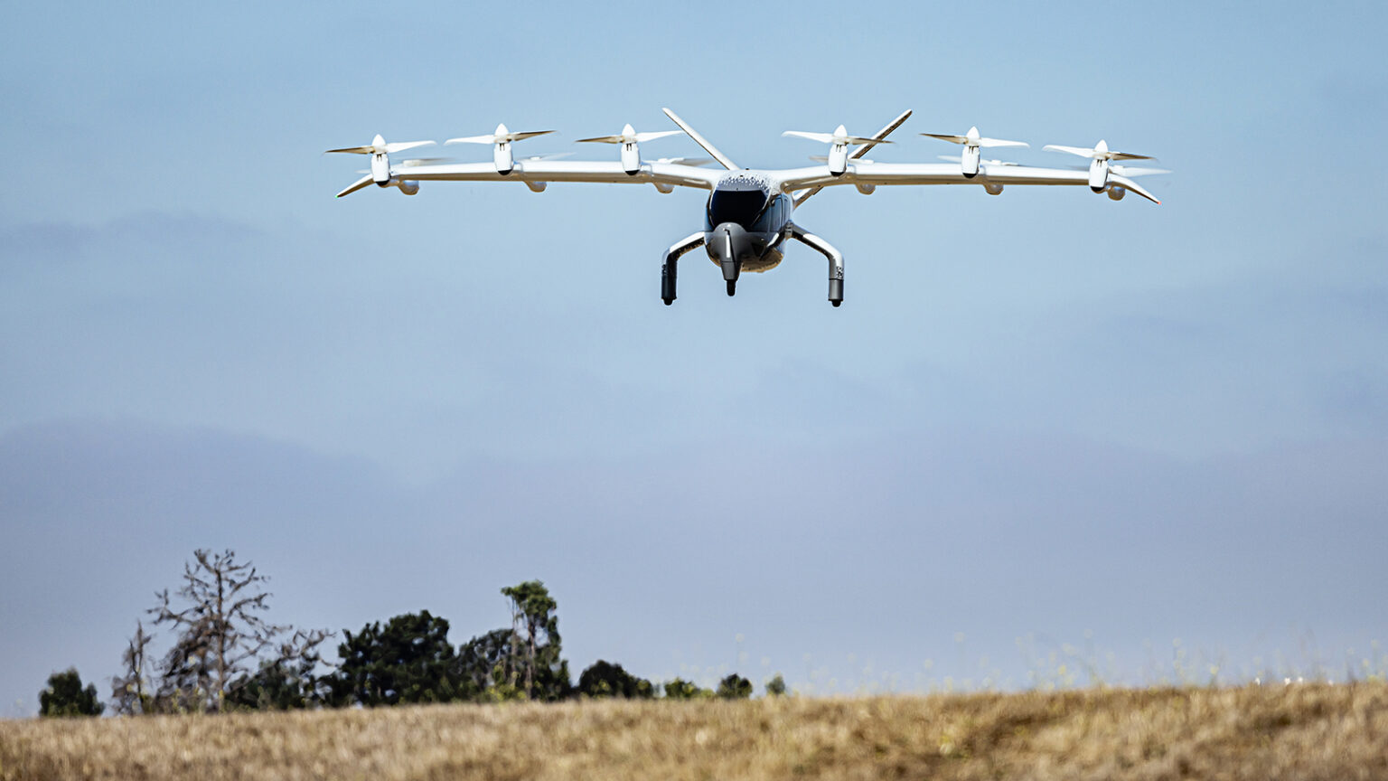 A white multicopter drone hovers in the sky over a grassy field with trees in the background.