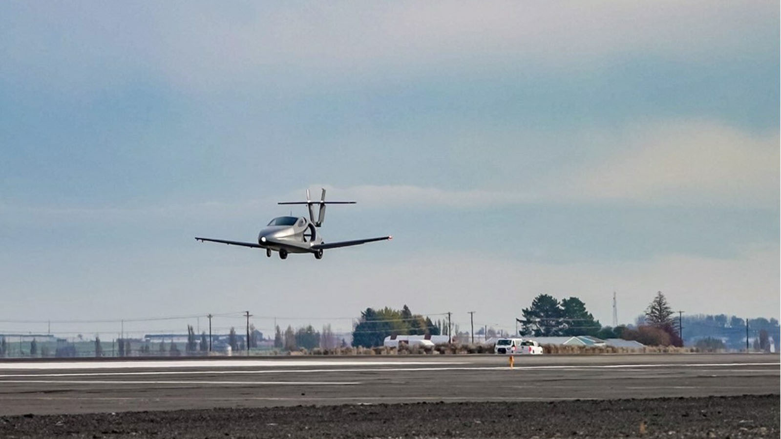 A small propeller aircraft in mid-air approaches a runway for landing, with a backdrop of a cloudy sky and sparse trees in the distance.