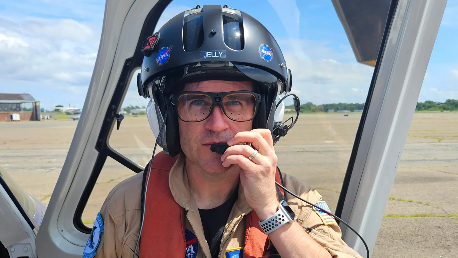 A person wearing a flight helmet and glasses, with a headset microphone, is inside a cockpit. The helmet has patches and the word "Jelly." The background shows an airstrip and partly cloudy sky.