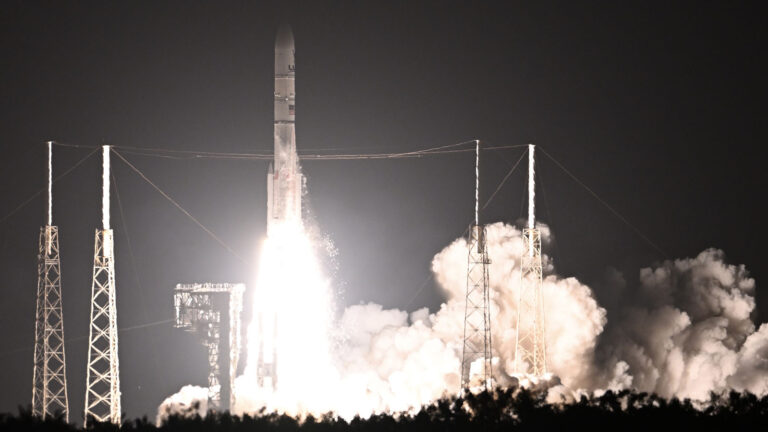 A rocket launches into the night sky with bright flames and smoke, surrounded by support structures.
