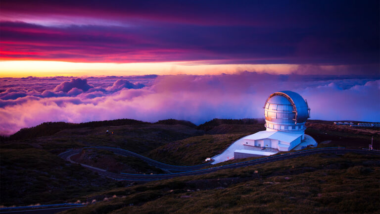A large observatory dome sits atop a hill at sunset, surrounded by clouds and rugged terrain, with winding roads leading to it.
