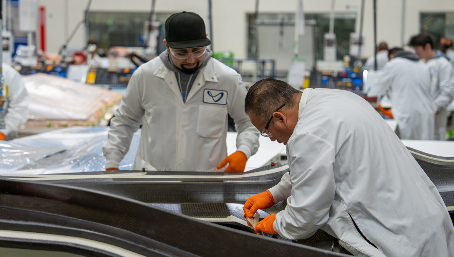 Two workers in white lab coats and gloves assemble materials in a factory setting. Plastic sheets and industrial equipment are visible in the background.