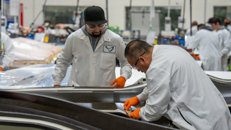 Two workers in white lab coats and gloves assemble materials in a factory setting. Plastic sheets and industrial equipment are visible in the background.