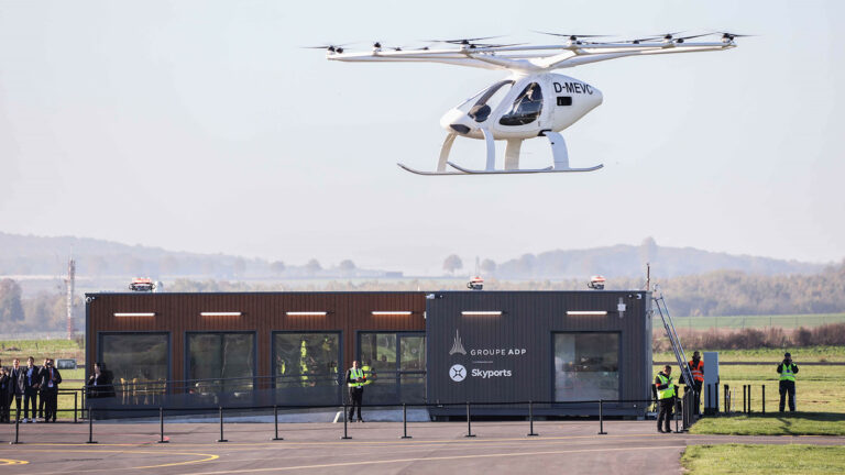 A white drone helicopter hovers above a small building labeled "Groupe ADP Skyports." Several people in high-visibility vests stand nearby, observing the flight.