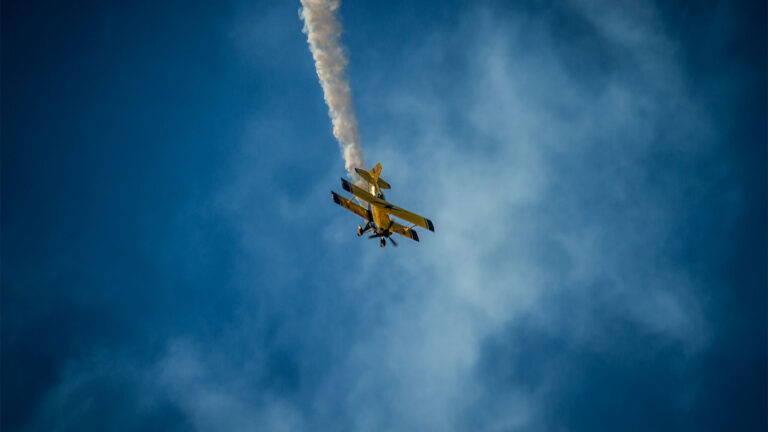 A yellow biplane emits white smoke while performing an aerial maneuver against a blue sky.
