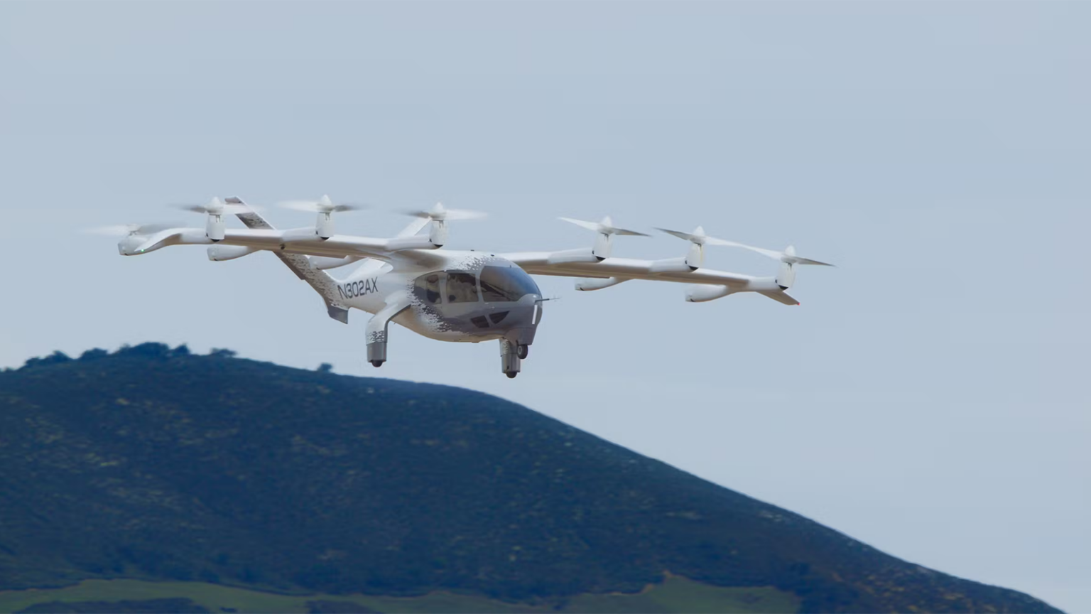 A white quadcopter drone in flight against a background of hills and a clear sky.