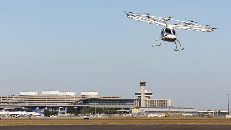 A multirotor aircraft hovers near an airport with a large terminal building and control tower in the background.