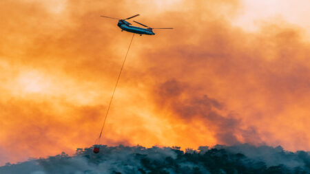 A helicopter carries a bucket of water above a forest fire, with thick smoke and flames below.