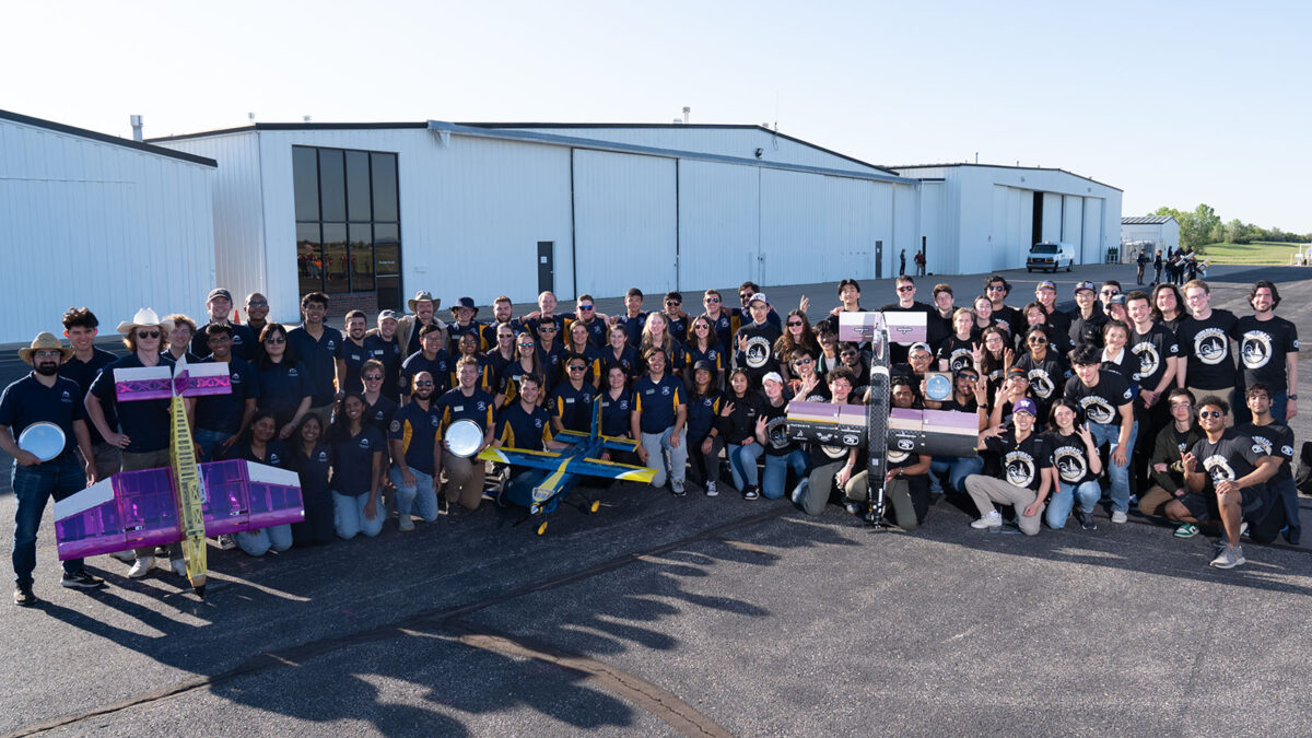 A large group of people posing outdoors in front of a few hangars, some holding model airplanes and team awards.