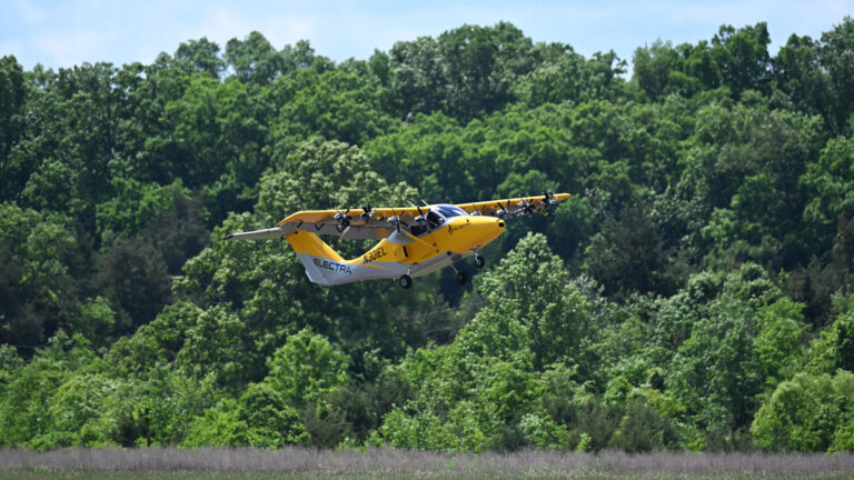 A yellow and white electric airplane is in flight over a grassy area with dense green trees in the background.