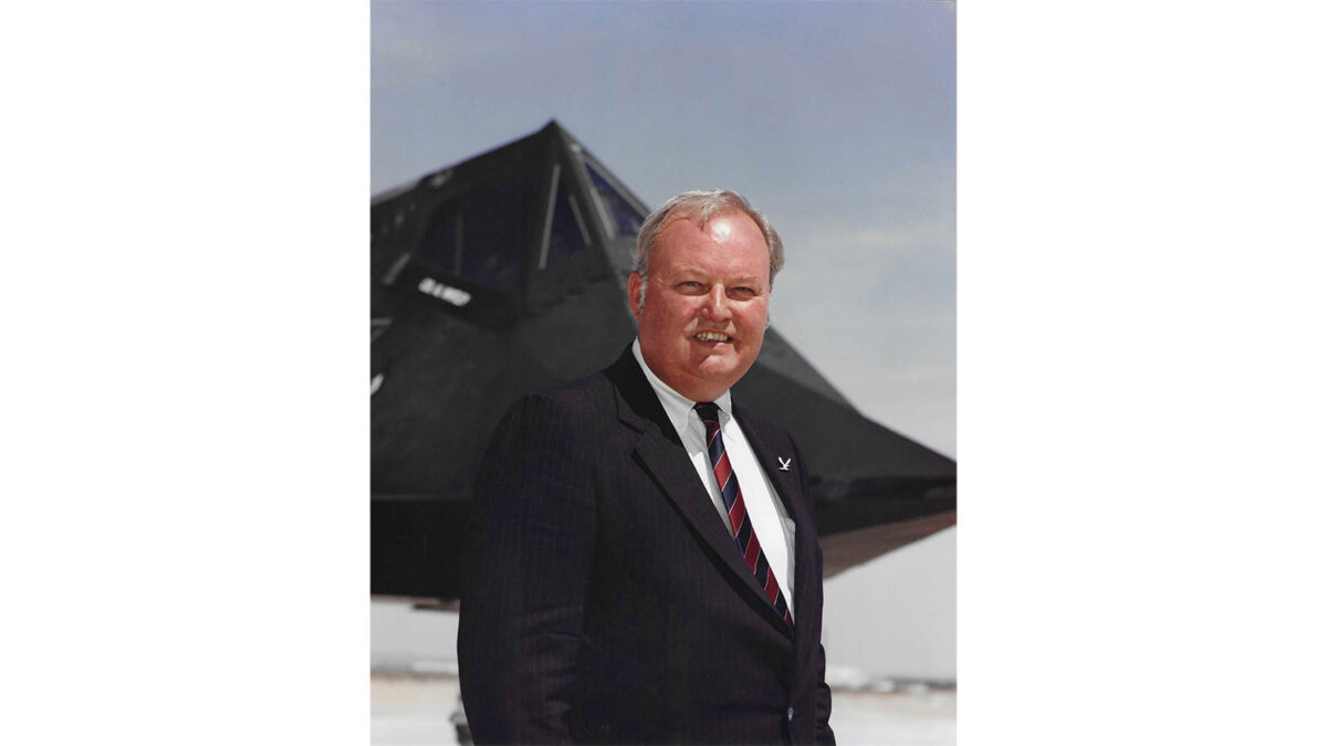A man in a suit and tie stands in front of a black stealth aircraft, smiling at the camera against a clear sky background.