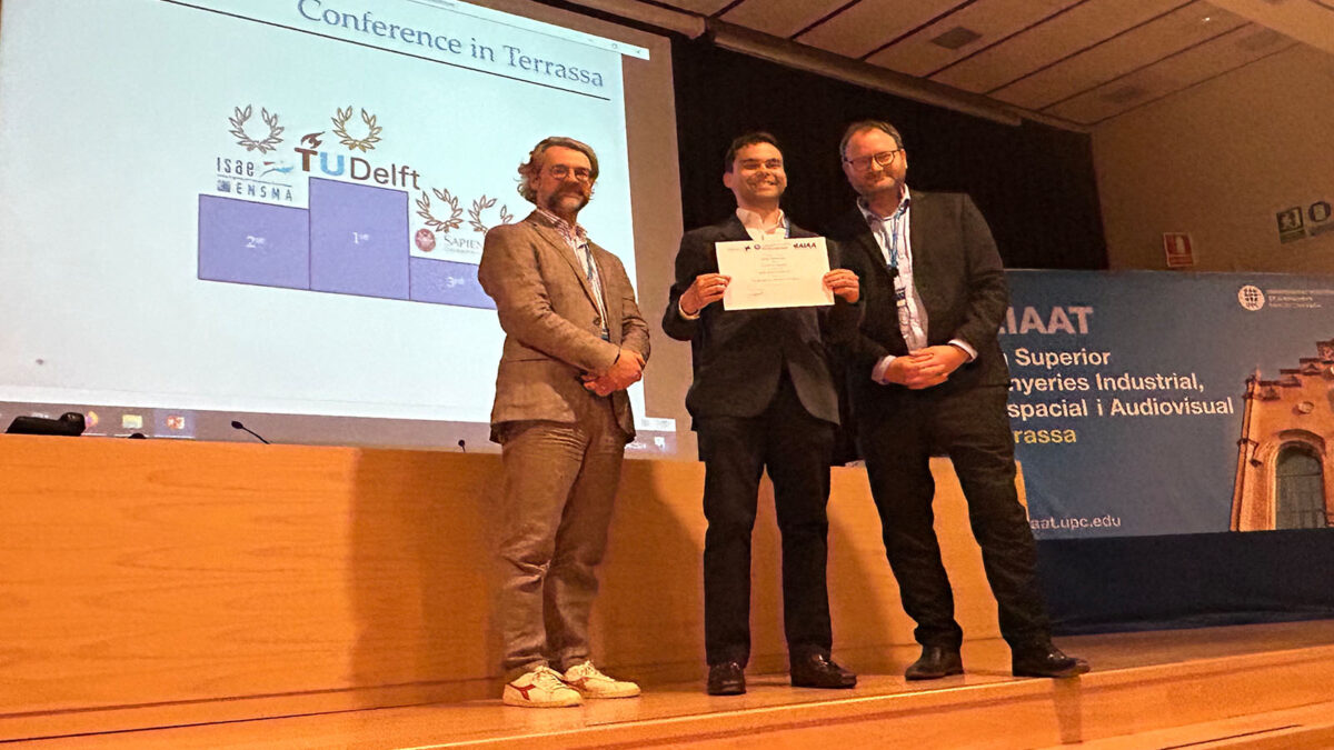 Three men in formal attire stand on a stage, one holding a certificate, during a conference in Terrassa, with a presentation slide and a blue banner in the background.