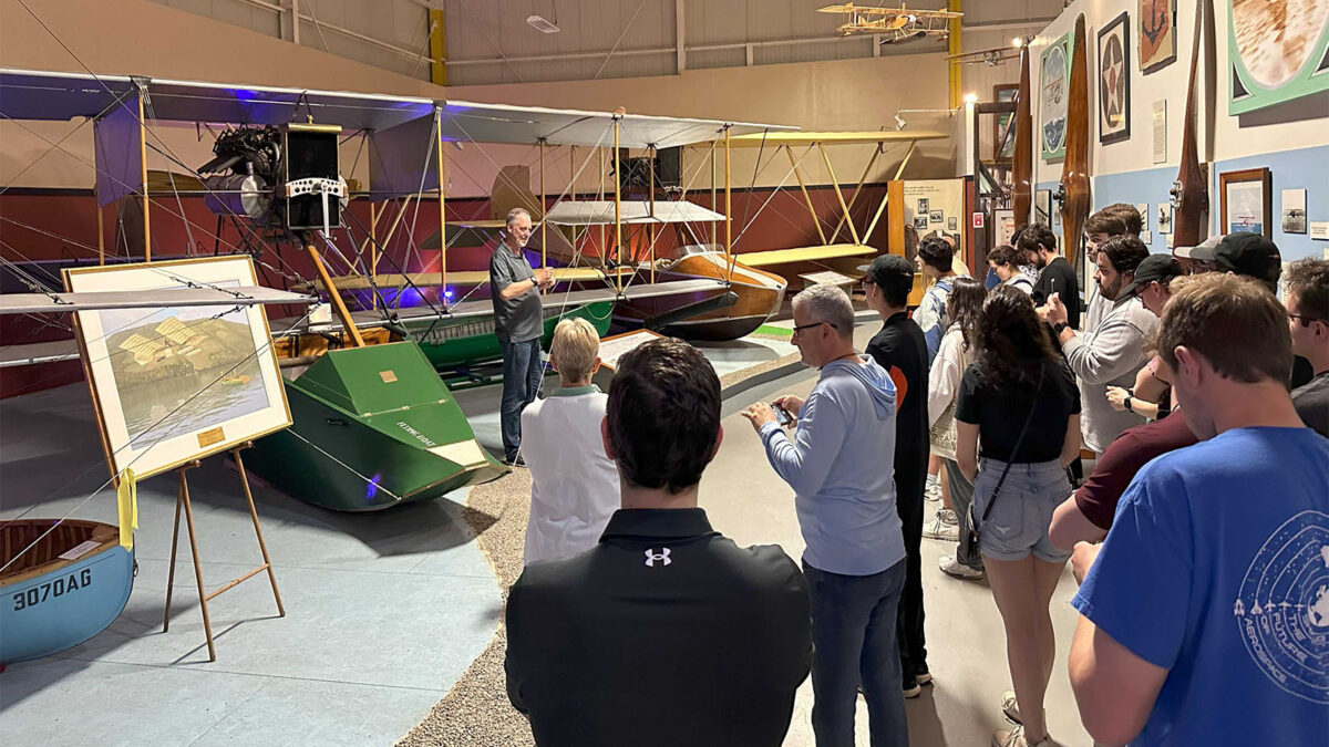 A group of people is gathered in a museum or exhibition hall listening to a person giving a presentation about vintage aircraft, with several airplanes displayed around them.