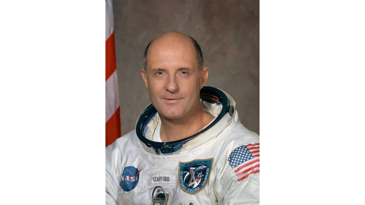 A bald astronaut wearing a NASA spacesuit poses for a formal portrait. An American flag is partially visible in the background.