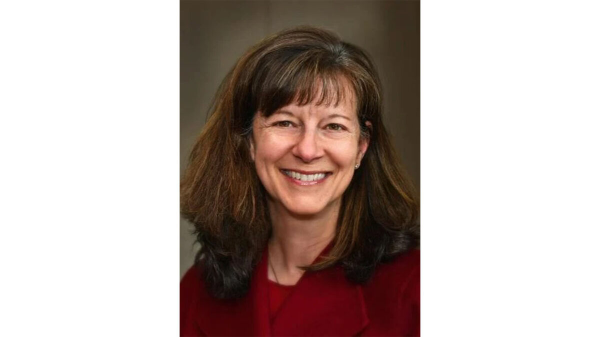 A woman with shoulder-length brown hair and a fringe smiles at the camera, wearing a maroon top and a neutral background.