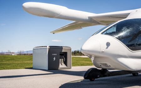 A white electric aircraft is parked on a pavement near a charging station labeled "CHARGE" with a lightning bolt symbol. Green grass and a clear blue sky are in the background.