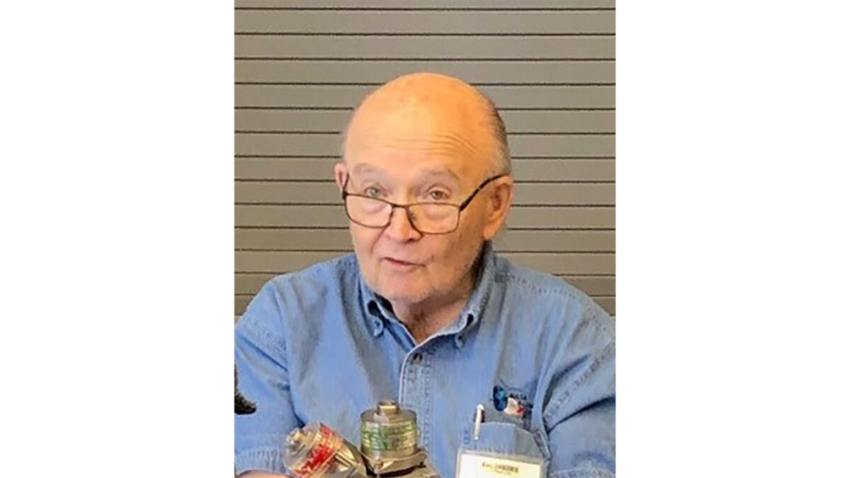 An older man with glasses in a blue shirt sits in front of a gray background, speaking with a microphone and various equipment in front of him.