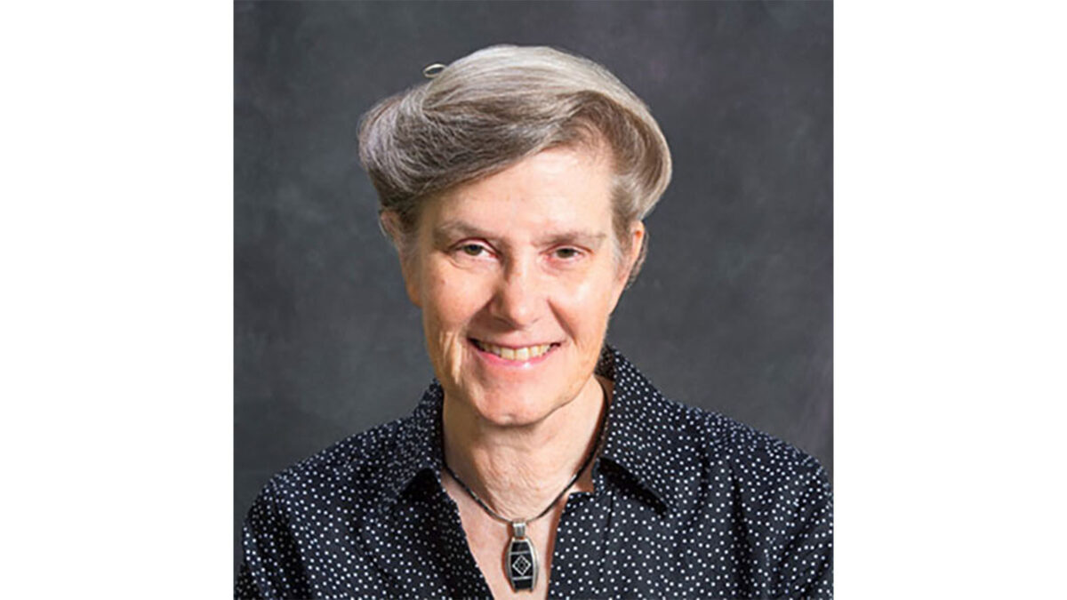 A person with short, gray hair is smiling at the camera. They are wearing a black, polka-dotted shirt and a pendant necklace. The background is dark gray.