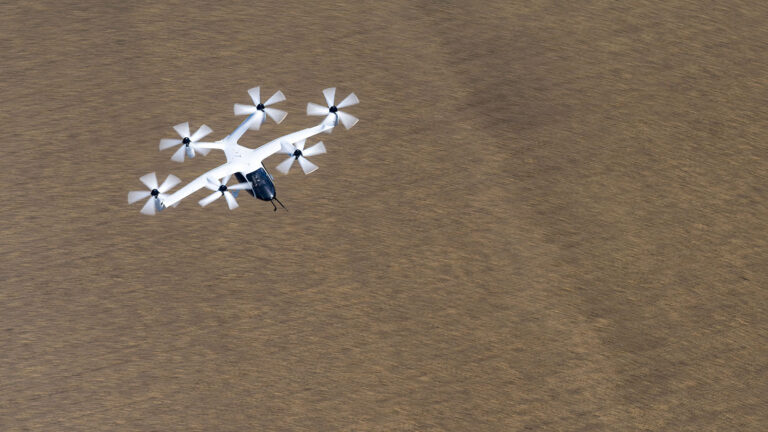 A white and black drone with six propellers flies over a brown, barren landscape.